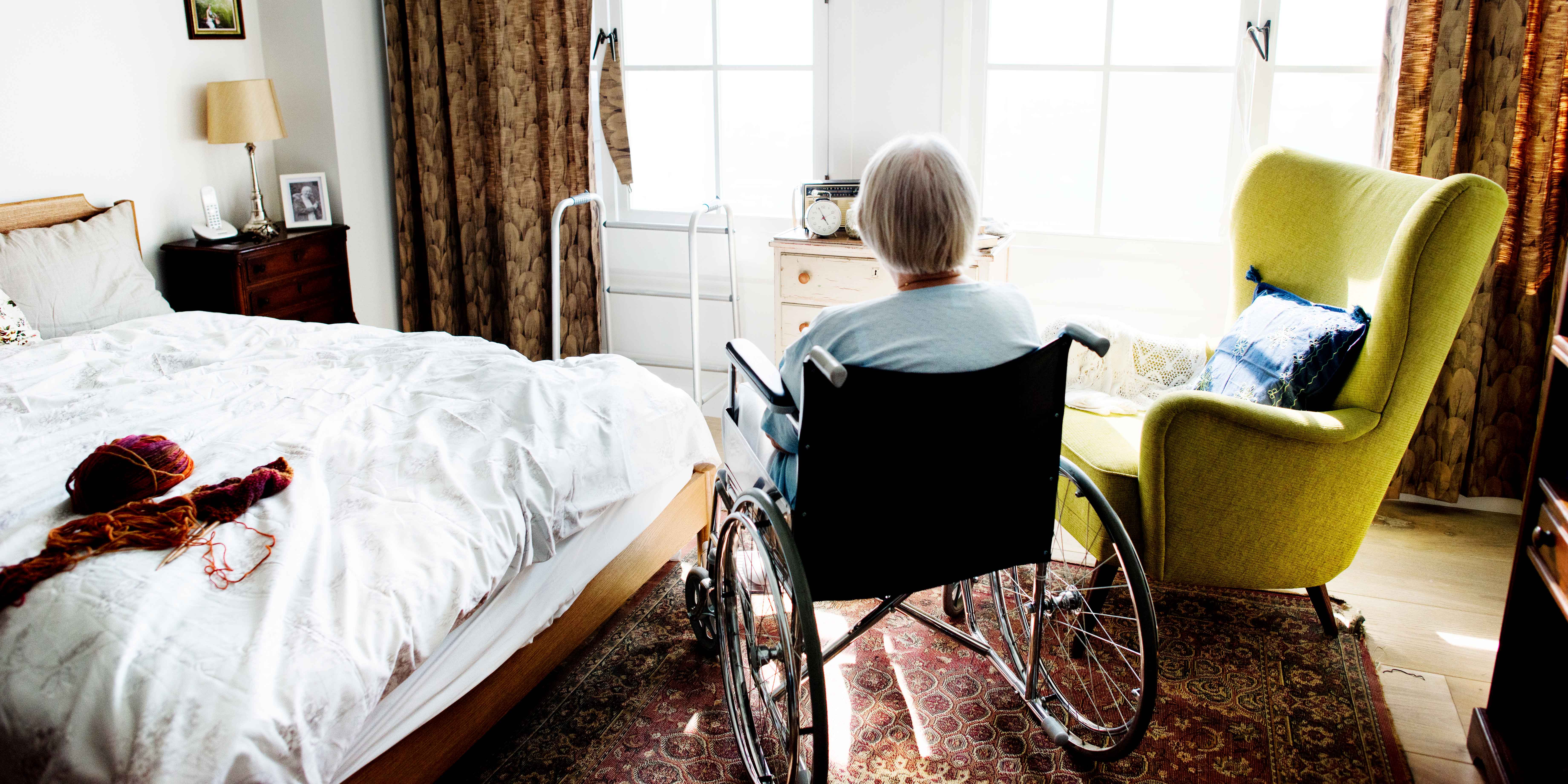 A woman with dementia sits in her bedroom at home looking out a window.