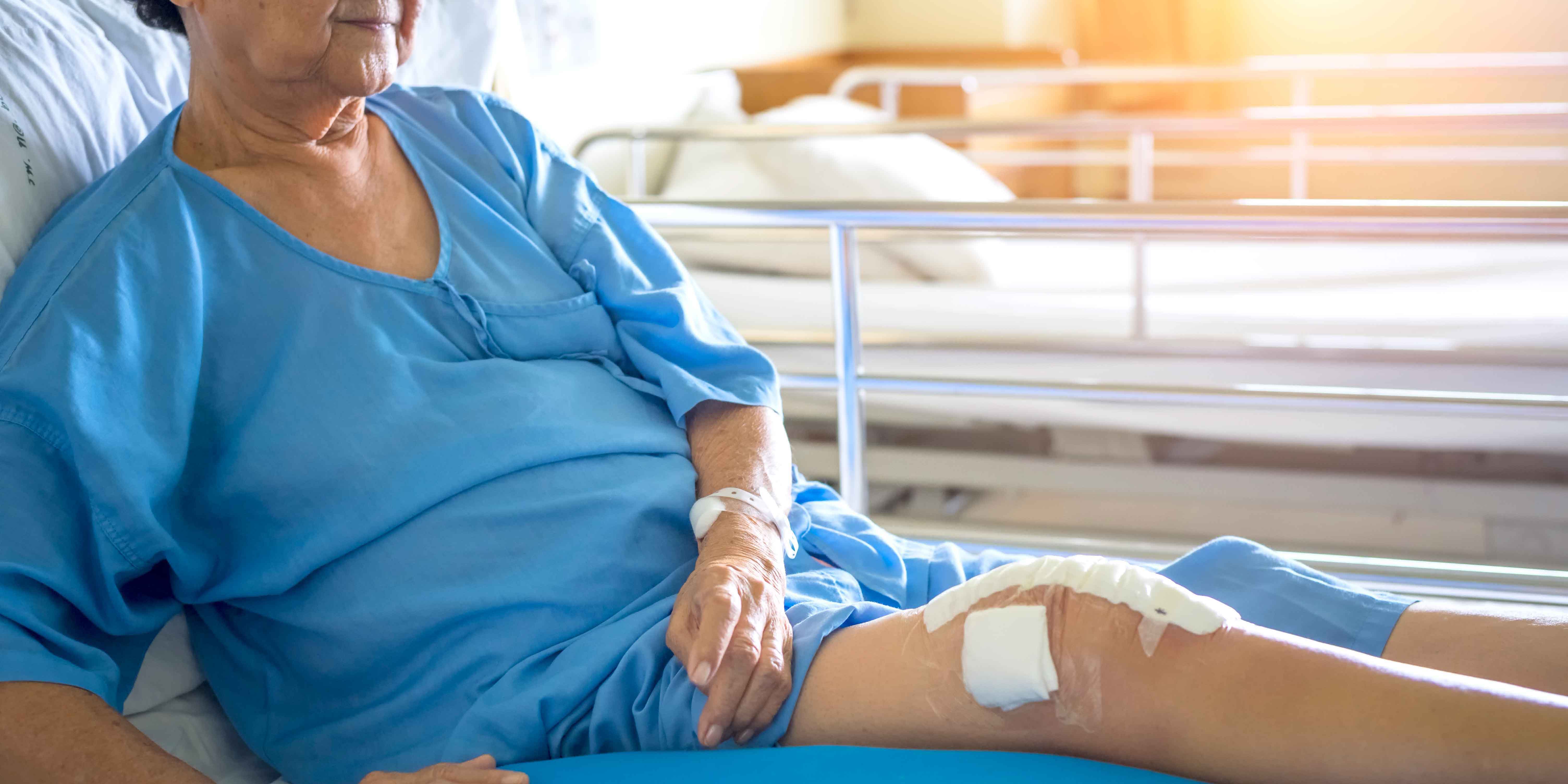 An elderly female patient sits on her bed with her knee wounds bandaged.