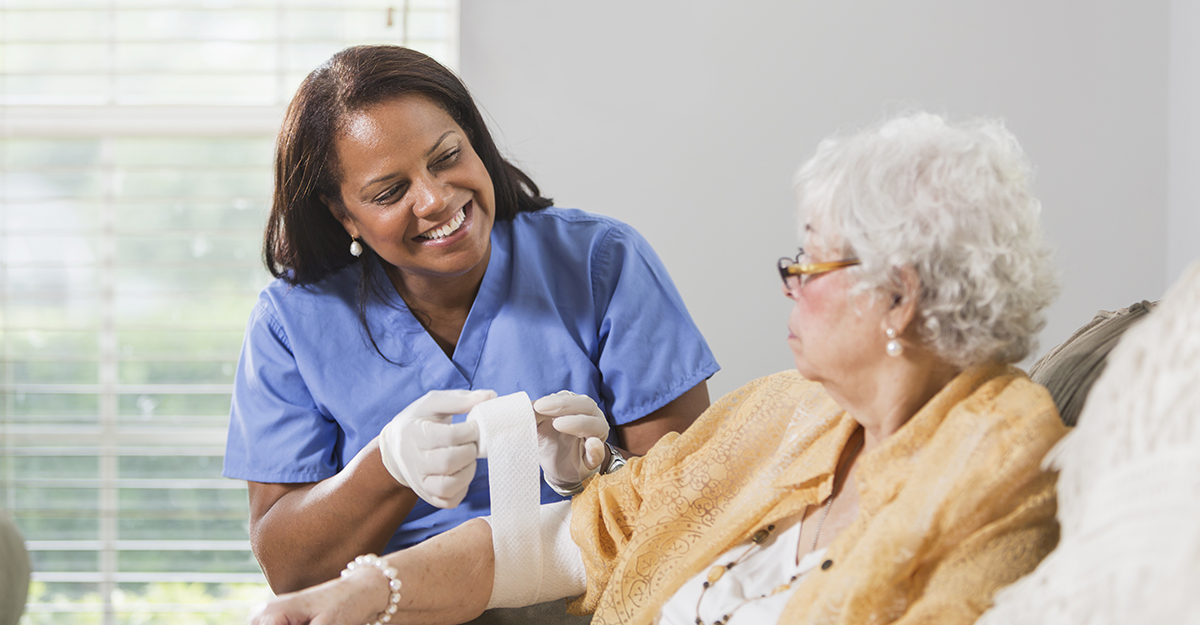 Senior Hispanic woman (70s) with Hispanic healthcare worker (50s) wrapping arm in bandage. Focus on healthcare worker.